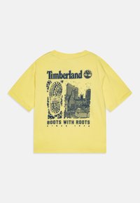 Dzeltena kokvilnas T-kreklu ar īsām piedurknēm, kas rotāta ar tumši zilu grafiku, kurā attēlots pilsētas siluets, zābaku pēda un uzraksts "Timberland Boots with Roots."