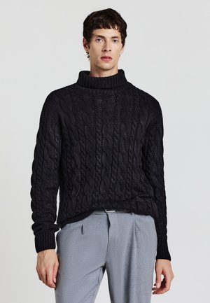 Pull en maille noire avec un col montant côtelé. Tissu épais et texturé avec une coupe ample. Associé à un pantalon gris clair.