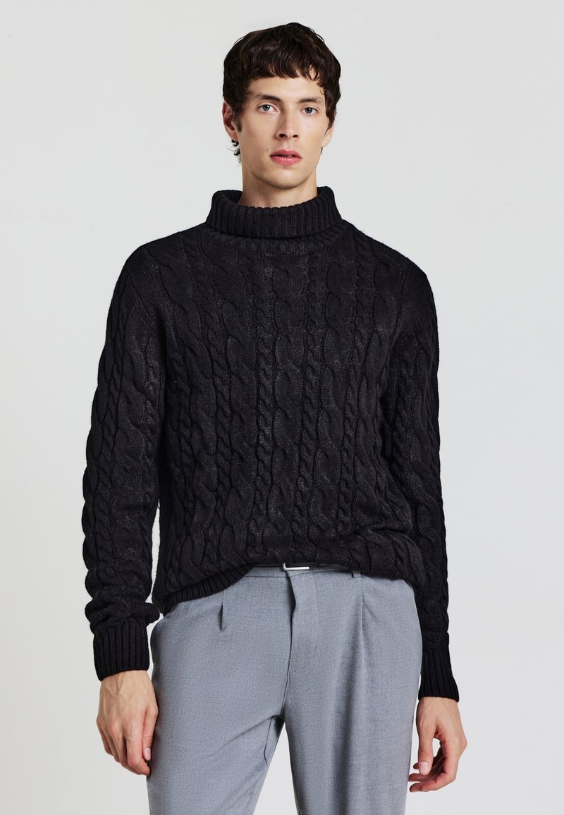 Maglione nero a maglia cordonata con colletto alto e a costine. Tessuto spesso e texture, con una vestibilità ampia. Abbinato a pantaloni grigi chiari.