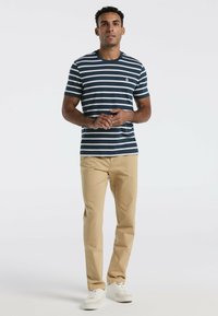 Homme debout portant une chemise à rayures bleu marine et blanche à manches courtes, un pantalon beige et des baskets blanches, contre un fond uni clair.