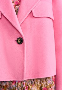 Chaqueta corta de lana rosa con solapa, que presenta un botón negro y dos bolsillos frontales. La tela tiene una textura suave.