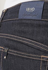 Vaqueros de mezclilla oscura con una superficie texturizada, que presentan costuras doradas, una etiqueta de parche de cuero azul y una pequeña etiqueta de marca cerca del bolsillo.