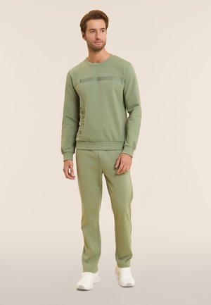 Man wearing olive green sweatshirt and matching pants with white sneakers, standing against a light beige background.