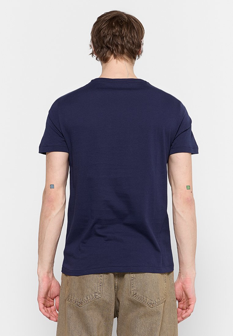 Uomo girato di spalle, indossando una semplice maglietta blu navy a maniche corte e jeans marrone chiaro, in piedi davanti a uno sfondo bianco semplice.