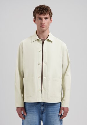 Ung mand iført en lys beige jakke med knapper og to forlommer over en brun skjorte og blå jeans, stående mod en ensfarvet baggrund.