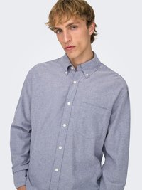 Lichtblauw overhemd van zachte stof; heeft lange mouwen, een kraag en een borstzak met witte knopen.