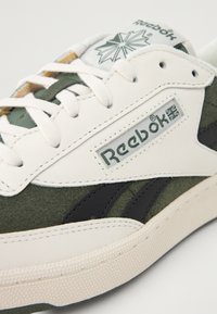 Reebok sneaker featuring a white leather upper, green suede accents, black lateral stripes, and textured rubber sole. Notable logo on the side.