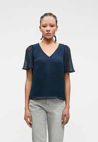 Σκούρο μπλε μπλούζα με κοντά διαφανή μανίκια, σχέδιο V-neck, απαλή υφή και άνετη εφαρμογή. Συνδυάζεται με ανοιχτό γκρι παντελόνι.