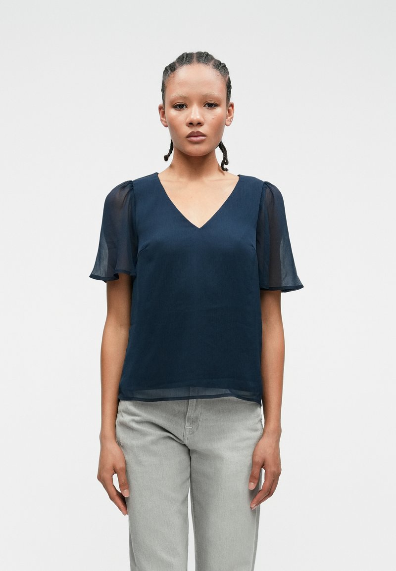 Σκούρο μπλε μπλούζα με κοντά διαφανή μανίκια, σχέδιο V-neck, απαλή υφή και άνετη εφαρμογή. Συνδυάζεται με ανοιχτό γκρι παντελόνι.