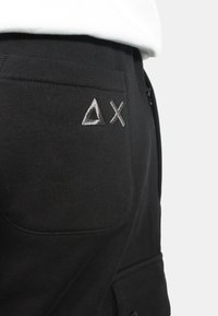 Pantaloni sportivi neri con una tasca posteriore decorata con forme a triangolo e X in ricamo argento. Il materiale ha una texture liscia e elastica.