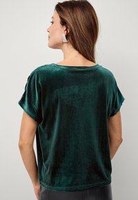 Grøn fløjl kortærmet top med løs pasform, rund halsudskæring og stikninger nær skulderen. Blød tekstur synlig.
