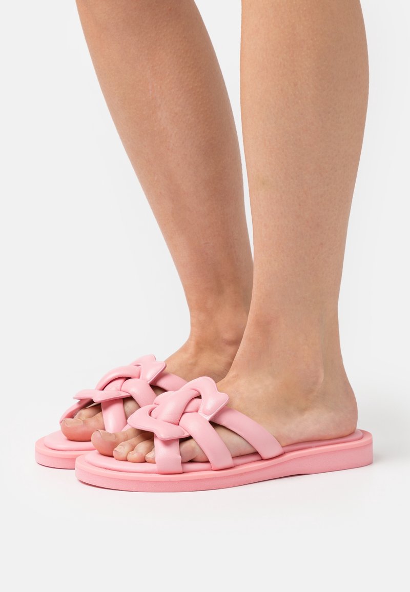 Pieds portant des sandales plates roses avec des brides épaisses et torsadées et un détail décoratif de nœud, présentés sur un fond clair uni.