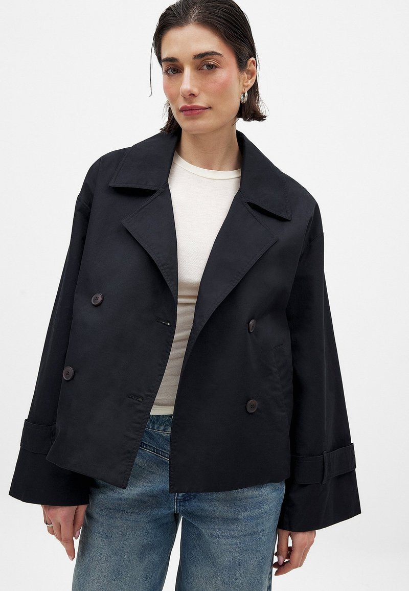 Svart dubbelknäppt jacka med bred krage, tillverkad av slätt tyg. Har stora knappar och lösa, korta ärmar. Bärs över en ljus skjorta.