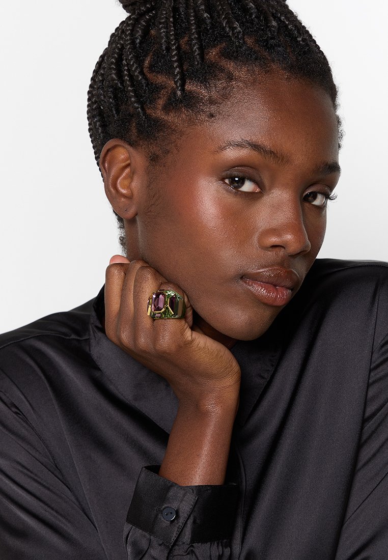 Bague dorée avec une grande pierre précieuse violette et verte, sertie dans une bande texturée, portée sur une main contre une chemise en satin noir.