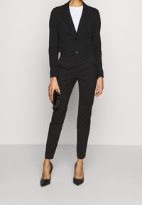 Femme portant un blazer noir ajusté et court, un pantalon noir fuselé, des talons noirs pointus, un haut blanc, tenant une petite pochette noire dans la main droite.