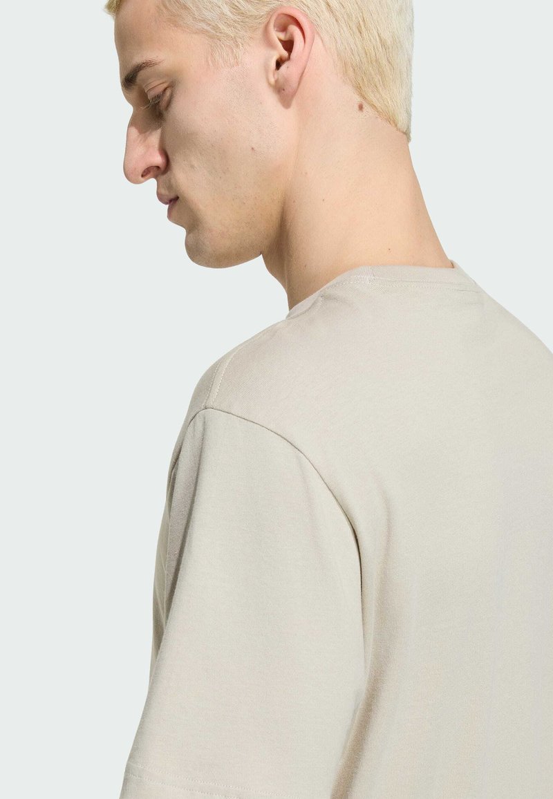 Jonge man met kort gebleekt haar die een lichtbeige T-shirt draagt, zijwaarts kijkend met ogen dicht tegen een effen achtergrond.