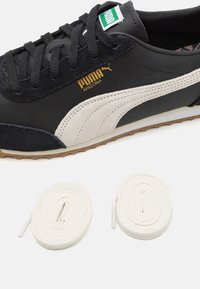 Svart och beige Puma-sneaker med överdel i slätt läder, mockadetaljer och texturerad yttersula; innehåller två vita skosnören.