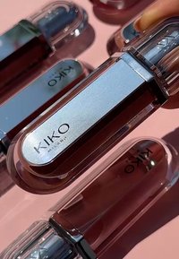 KIKO Milano lip products in transparent plastic tubes with silver accents. Various shades of brown and nude are visible against a pink background.