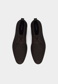 Σκούρα καφέ δερμάτινα ανδρικά επίσημα παπούτσια με κορδόνια, κορυφαία όψη, που δείχνει στρογγυλεμένες μύτες και θηλιές έλξης στις φτέρνες σε απλό φόντο.