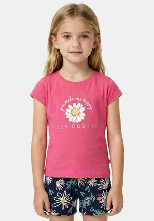 Jeune fille aux cheveux blonds portant un t-shirt rose avec une marguerite et le texte "Tu me rends heureux" ainsi qu'un short bleu marine à motif floral de marguerites.