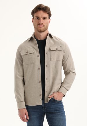 Camisa abotonada de color beige claro con dos bolsillos en el pecho, cuello y botones marrones. Lleva puesta sobre una camiseta negra de cuello redondo, combinada con vaqueros azules.