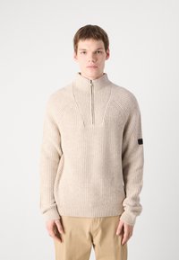 Beige bordázott kötött pulóver magas nyakkal és fél cipzárral. Szögletes nyakú kialakítással és testhezálló mandzsettákkal rendelkezik. Diszkrét logó folt az ujjon.
