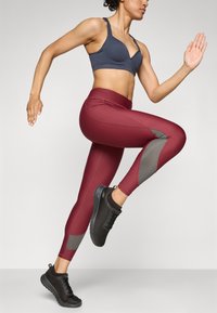 Bordo atletske tajice s sivim mrežastim detaljima i visokim strukom, u paru s tamnosivim sportskim grudnjakom i crnim tenisicama za trčanje.