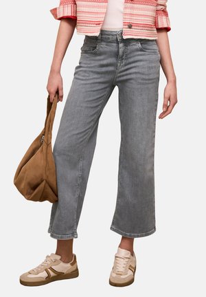 Kobieta ubrana w szare jeansy o szerokich nogawkach, beżowe trampki oraz różową kurtkę w wzory, trzymająca w dłoni brązową miękką torebkę.