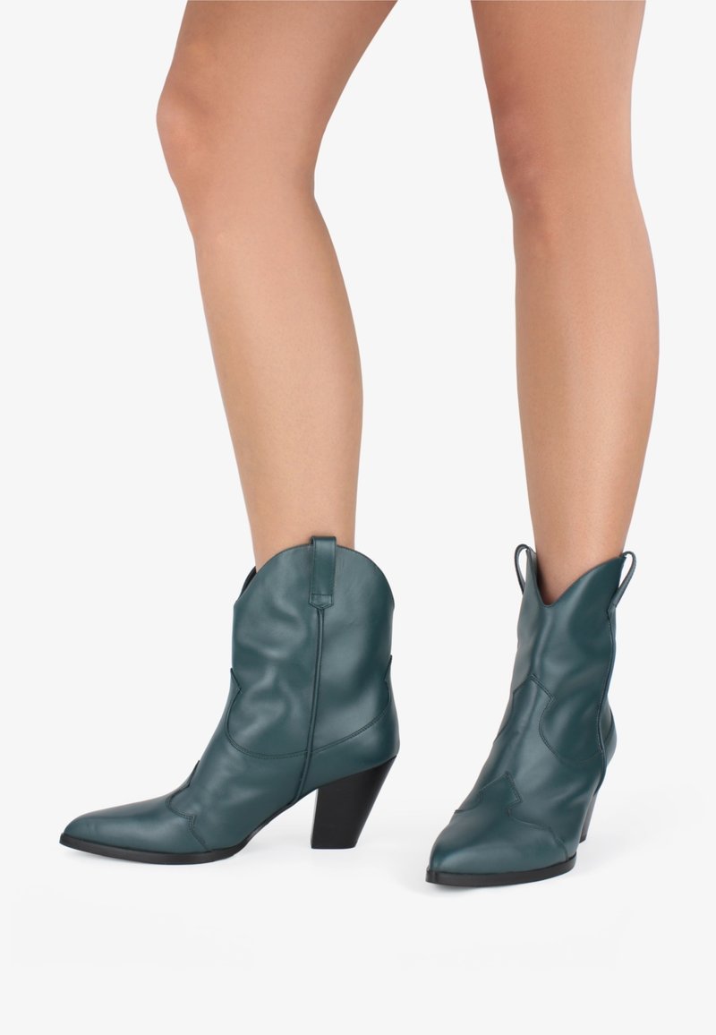 Botas de vaquero de cuero verde azulado con punteras puntiagudas y tacón alto. Presenta costuras al estilo western y tiradores para facilitar su calzado. Textura suave.