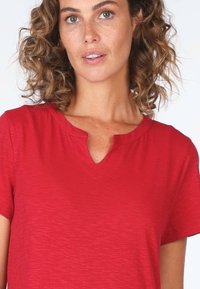 Rotes T-Shirt mit kurzen Ärmeln aus strukturiertem Stoff und V-Ausschnitt. Einfaches Design, betont natürliche Kurven mit lockerer Passform.