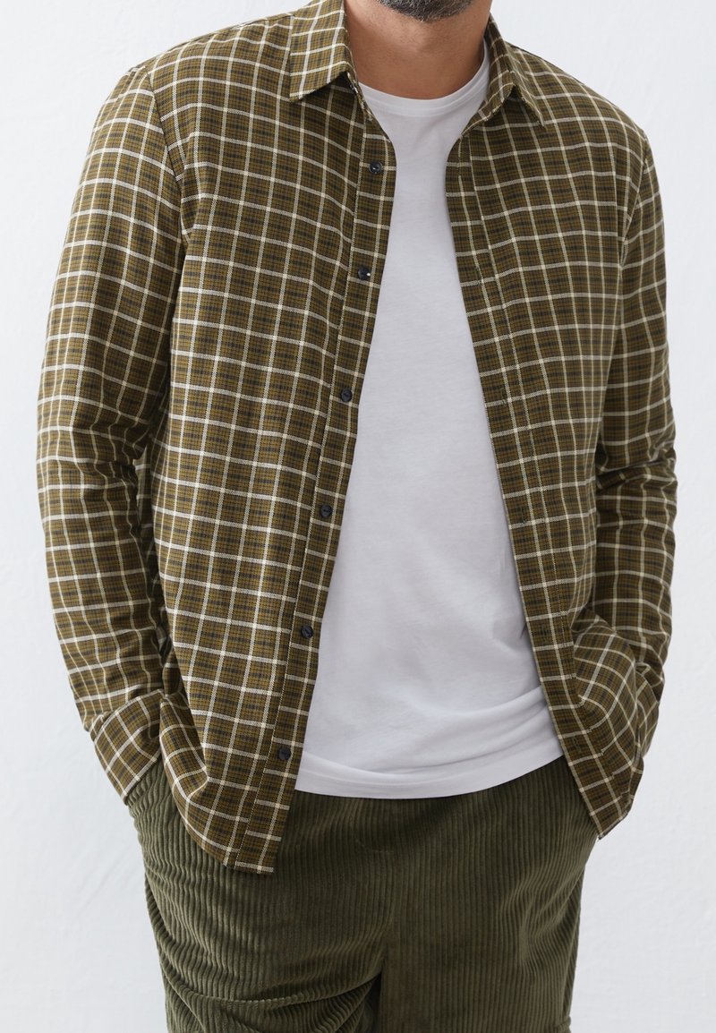 Groen-bruine geruite blouse met lange mouwen, knopen aan de voorkant en een kraag, gedragen over een wit T-shirt; gecombineerd met olijfgroene ribfluwelen broek.