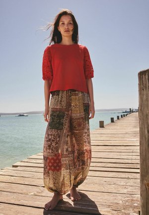 Žena v červenej blúzke a vzorovaných širokých nohaviciach stojaca bosá na drevenom móle pri mori pod jasnou oblohou.