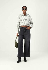 Cropped white shirt with black abstract print, high-waisted dark denim wide-leg pants, black ankle boots, and green shoulder bag.