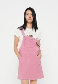 Rosa Corduroy-Latzkleid mit verstellbaren Trägern, aufgesetzter Fronttasche und seitlichen Taschen, getragen über einem weißen T-Shirt mit Grafikdruck.