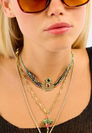 Colliers superposés en ton or avec des accents de perles. Comprend un pendentif en forme d'oiseau avec des pierres noires et rouges. Lunettes de soleil à verres bruns.