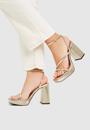 Metallic gold high-heeled sandals with double front straps, ankle strap, and a chunky square heel; smooth finish and open toe design.