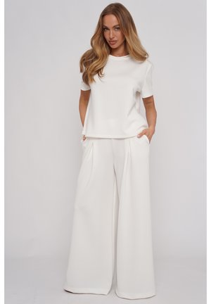 Kobieta z długimi falowanymi włosami, ubrana w biały krótki top i białe szerokie spodnie, stoi ze rękami w kieszeniach na jednolitym tle.