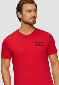 Rode katoenen t-shirt met korte mouwen, met zwarte tekst: "FISH 'N CHIPS Levering VERHIT bij jou thuis TEL 765-34-2184 FISH."