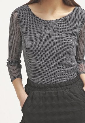 Frau trägt ein schwarz-weiß gemustertes langärmeliges Oberteil mit transparenten Ärmeln und schwarze, hoch taillierte gesteppte Hosen, die Hände in den Taschen.