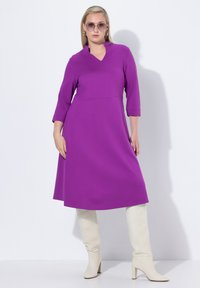 Robe violette avec un décolleté en V et des manches trois-quarts, en tissu lisse. Portée avec des bottes blanches tailles genou et de grandes lunettes de soleil.
