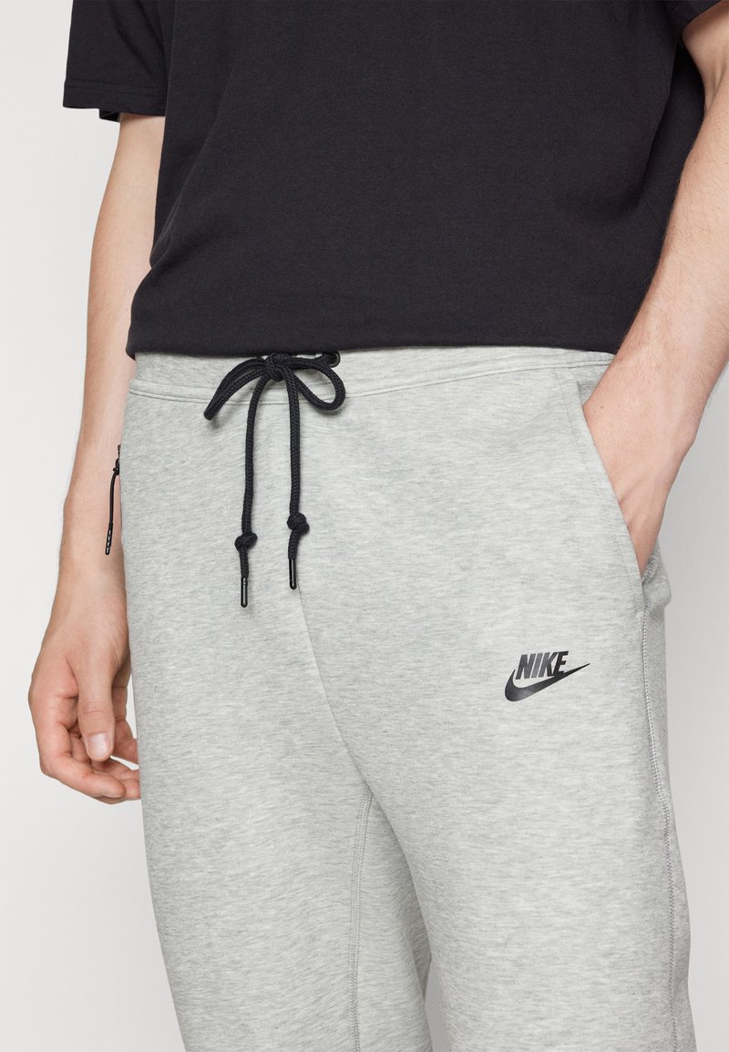 Graue Jogginghose mit einem Kordelzugbund, seitlichen Taschen und schwarzem Nike-Logo am linken Oberschenkel. Glatte Textur, lässiges Design.