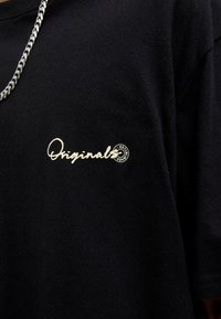 Černé bavlněné tričko s bílou vyšívanou značkou "Originals" a kruhovým detailem na levé části hrudi. Krátké rukávy, střih s kulatým výstřihem.