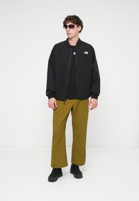 Μαύρο μπουφάν bomber με κλείσιμο με φερμουάρ και ελαστικά μανίκια, σε συνδυασμό με χακί παντελόνι και μαύρα παπούτσια. Αξεσουάρ περιλαμβάνουν σκουρόχρωμα γυαλιά ηλίου.