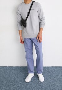 Hombre con sudadera ligera gris de Columbia, pantalones azul claro, zapatillas blancas y una mochila cruzada negra, de pie sobre una alfombra azul.