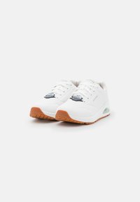 Zapatillas deportivas blancas con parte superior texturizada, suela de goma y suela exterior marrón. Sus características incluyen cordones y una almohadilla de aire visible.