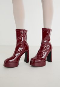 Burgunder patent lær ankle boots med en høj blokhæle, lynlås lukning og bølge-lignende design foran. Glat, blank tekstur.