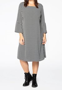 Černobílé šaty s kostkovaným vzorem, se čtvercovým výstřihem a zvonovými rukávy, vyrobené z měkké látky, doplněné černými šněrovacími kotníkovými botami.