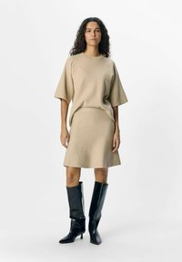 Beige striktrøje med brede korte ærmer parret med en matchende flagrende nederdel. Sorte knælange hæl støvler fuldender looket.