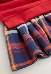 Rotes, geripptes Oberteil kombiniert mit einem marineblauen Karorock, der orangefarbene und weiße Akzente hat; weicher Stoff mit sichtbaren Falten am Saum.