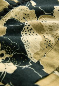 Tissu floral présentant de grandes fleurs beiges et noires, avec des accents vert foncé. Le textile montre une texture douce et des coutures détaillées.